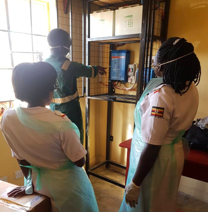 An electrician points at a panel on a shelf while two nurses observe. The room has yellow walls and sunlight is streaming in from the window on the left, creating a hazy, overexposed effect